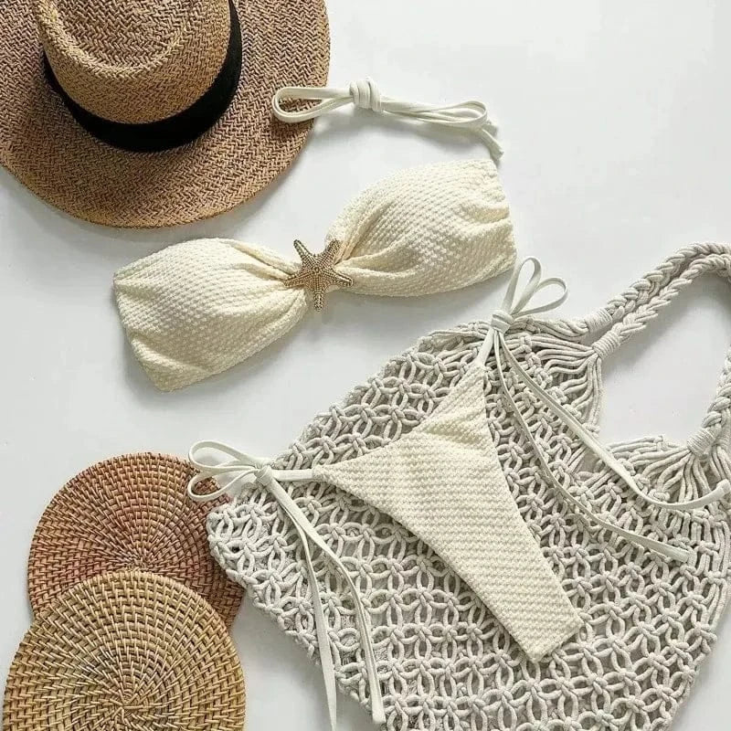 A white textured strapless bikini with a gold starfish accent, accompanied by a straw hat, two round woven mats, and a cream macrame tote bag on a white background.