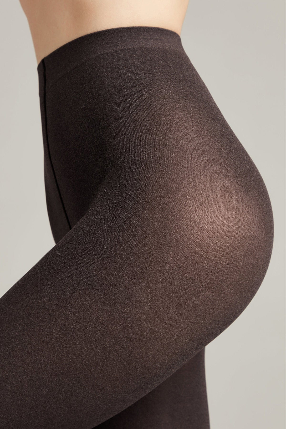 A close-up side view of dark brown, sheer tights worn on a person, featuring a high-waisted design against a plain light gray background.