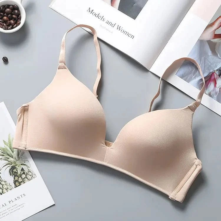 A beige wireless bra with adjustable thin straps and smooth molded cups displayed on a gray surface next to an open magazine and a small bowl of coffee beans.