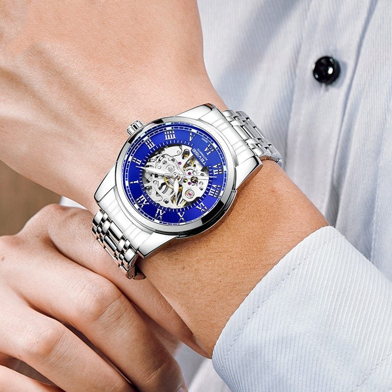 A silver-tone automatic watch with a blue circular dial, Roman numeral indices, and an exposed mechanical skeleton movement worn on a person's wrist.