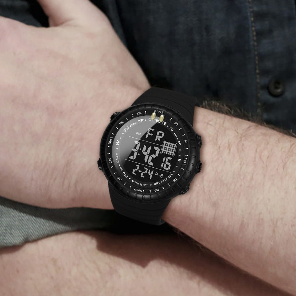 A black digital sports watch with a round face, compass markings on the bezel, and a silicone strap is worn on a person's wrist.