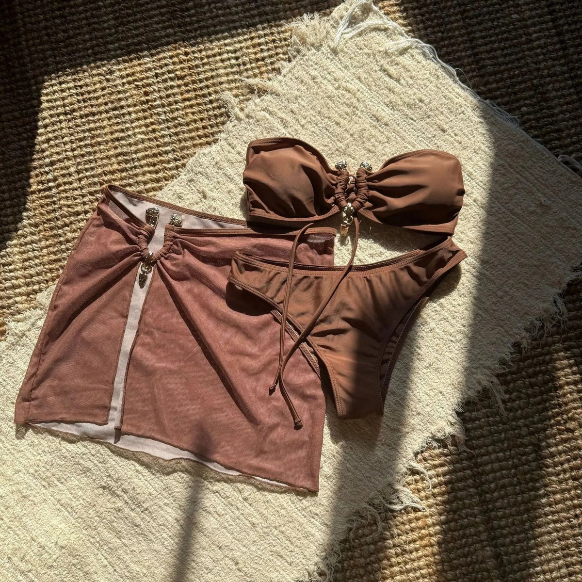 A brown strapless bikini set consisting of a gathered bandeau top with a central gold-tone ring detail, high-cut bottoms, and a matching sheer mesh mini skirt laid out on a textured beige rug.