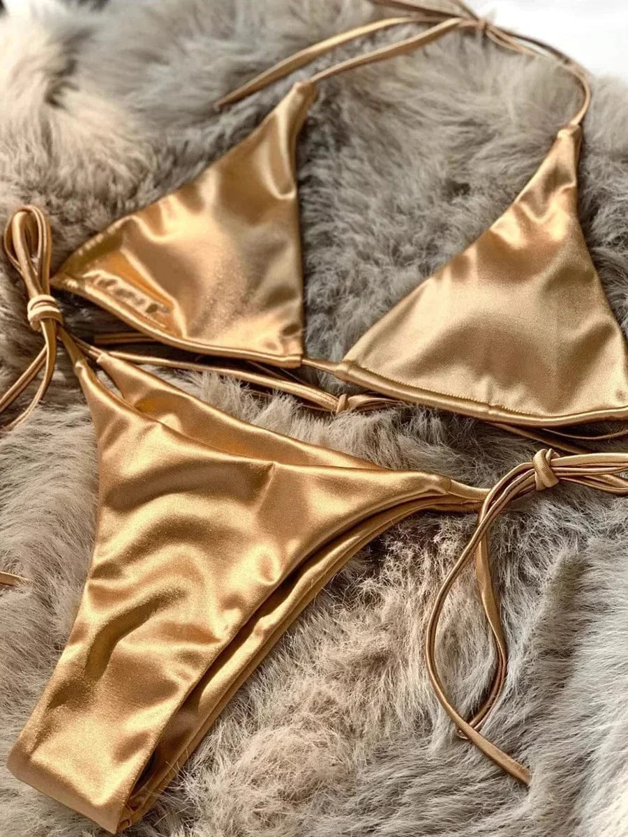A metallic gold bikini set featuring a string triangle top and matching tie-side bottoms, laid flat on a textured grey faux fur surface.