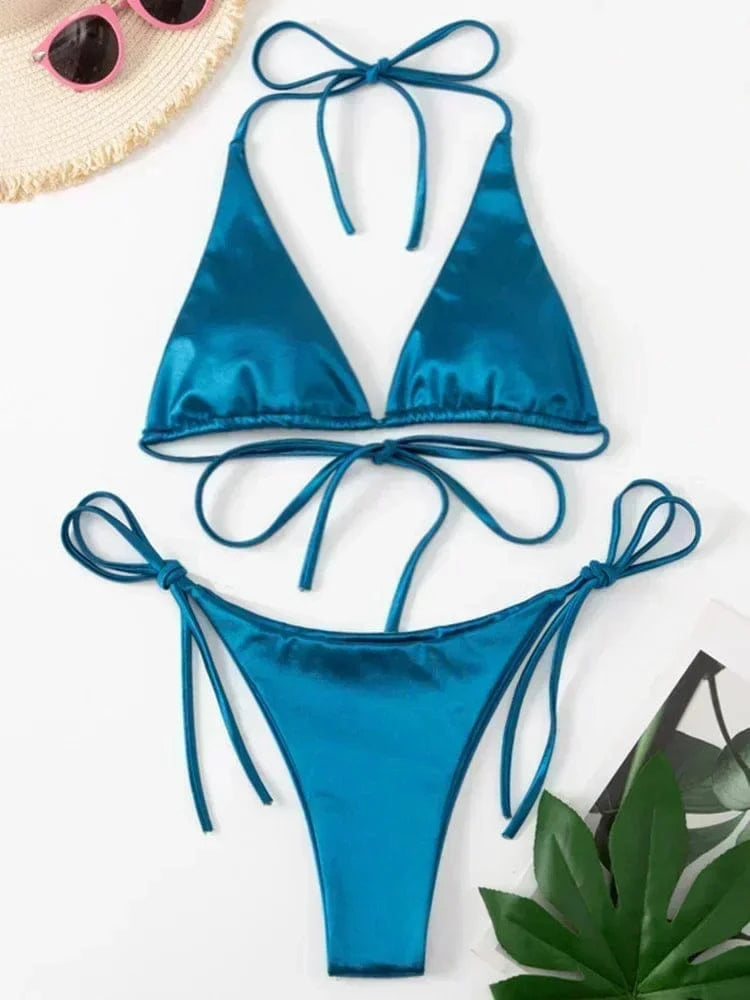 A metallic teal blue string bikini set with triangular cups and tie-side bottoms, laid flat on a white surface next to a straw hat with pink sunglasses and a green leaf.