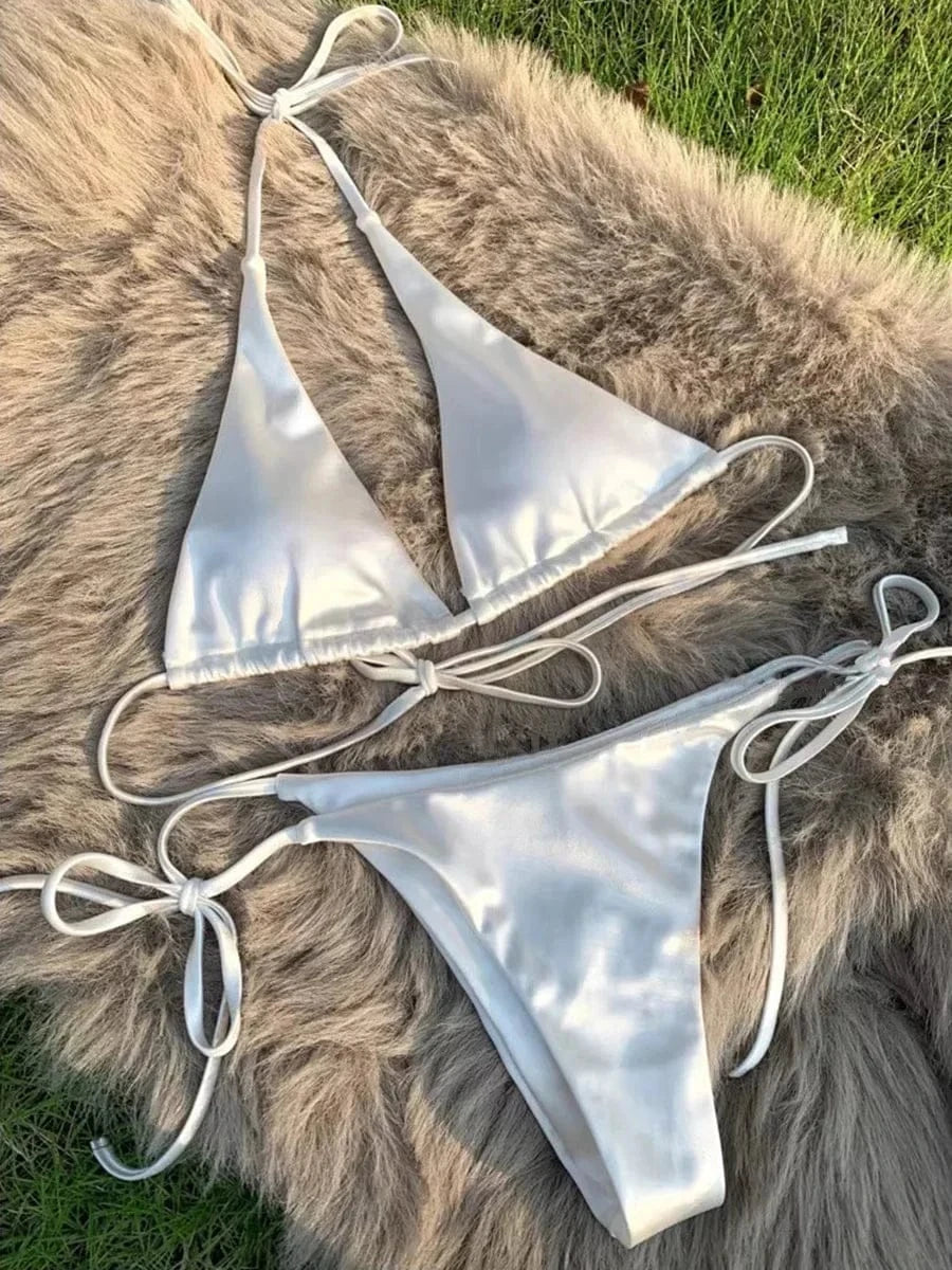 A white two-piece bikini set consisting of a triangle top with tie-neck straps and matching side-tie bikini bottoms, displayed on a fluffy beige rug.