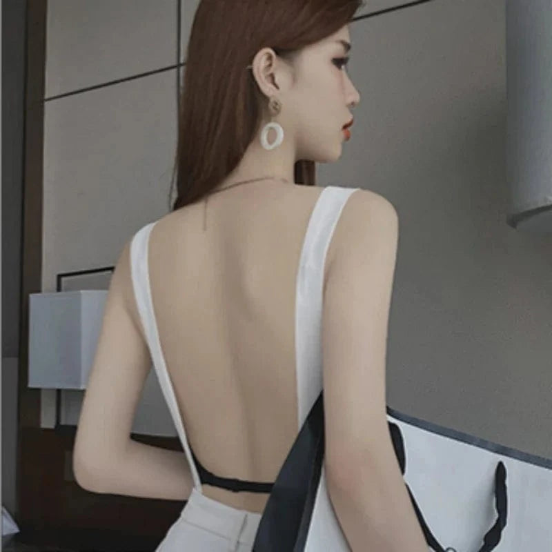 A woman wears a white backless tank top featuring a low-cut design with a black bra strap visible across her lower back, and carries a black and white tote bag.