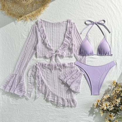 A light purple three-piece bikini set consisting of a triangle top, high-cut bottoms, a sheer long-sleeve tie-front cover-up top, and a matching ruffled skirt sarong laid flat on a white surface next to a sun hat and flowers.