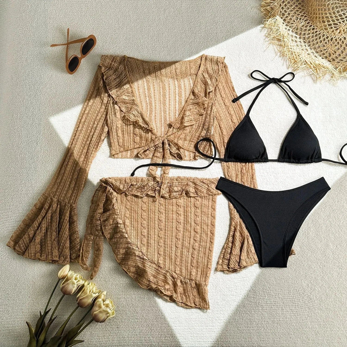A beige three-piece swimsuit set featuring a long-sleeved tie-front top and a ruffled sarong skirt, paired with a black ribbed triangle bikini set, with sunglasses and flowers placed on a textured white surface.