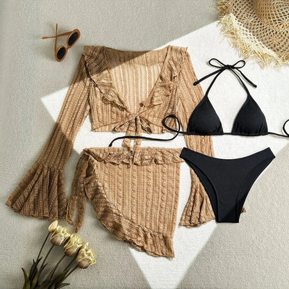 A beige three-piece swimsuit set featuring a long-sleeved tie-front top and a ruffled sarong skirt, paired with a black ribbed triangle bikini set, with sunglasses and flowers placed on a textured white surface.