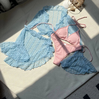 A light blue, long-sleeved, sheer lace bolero and matching mini skirt are laid flat next to a solid pink two-piece bikini on a cream-colored surface.