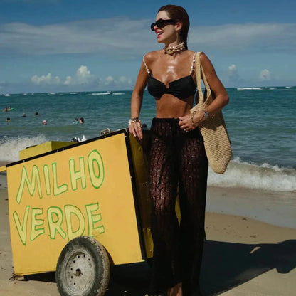 Black bikini top with pearl-accented straps and matching sheer, black crochet trousers worn by a woman posing on a sandy beach next to a yellow corn cart.