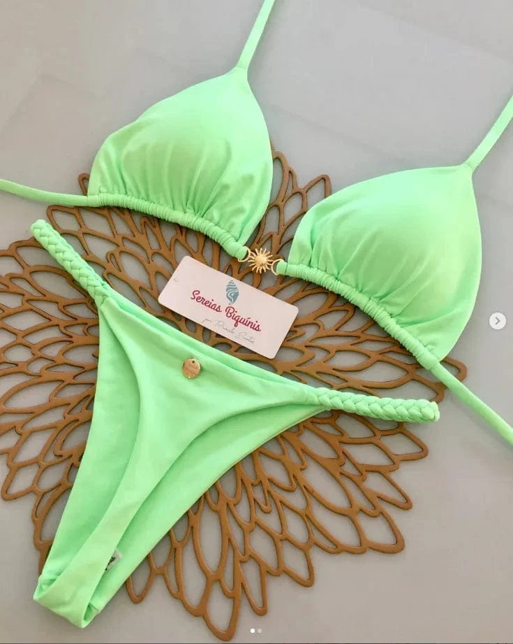 A light lime green bikini set featuring a triangle top with a central sun-shaped gold charm and a matching bottom with braided side straps resting on a decorative brown floral mat.