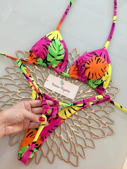 A colorful bikini set featuring a vibrant tropical leaf print in shades of hot pink, neon green, yellow, and orange, displayed on a decorative gold-toned placemat.