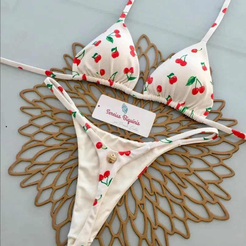 A white two-piece bikini featuring a red cherry pattern, displayed on a brown decorative placemat with a brand tag in the center.