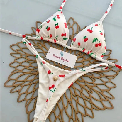 A white two-piece bikini featuring a red cherry pattern, displayed on a brown decorative placemat with a brand tag in the center.