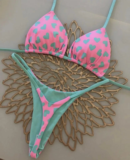 A two-piece bikini set featuring a pink triangle top and matching thong bottom, both patterned with light teal heart prints and accented with solid light teal straps, laid out on a gold-toned decorative placemat.