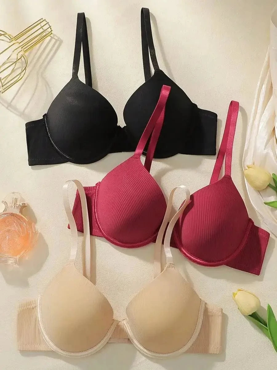 Three bras with ribbed fabric texture and adjustable straps, arranged in black, deep red, and beige on a light surface alongside a perfume bottle and two yellow tulip buds.