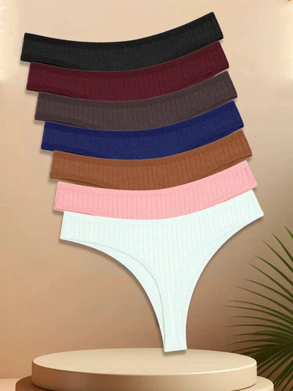 A stack of six ribbed thong underwear in black, dark red, dark brown, navy blue, light brown, soft pink, and white resting on a beige circular pedestal with a palm leaf visible in the bottom corner.