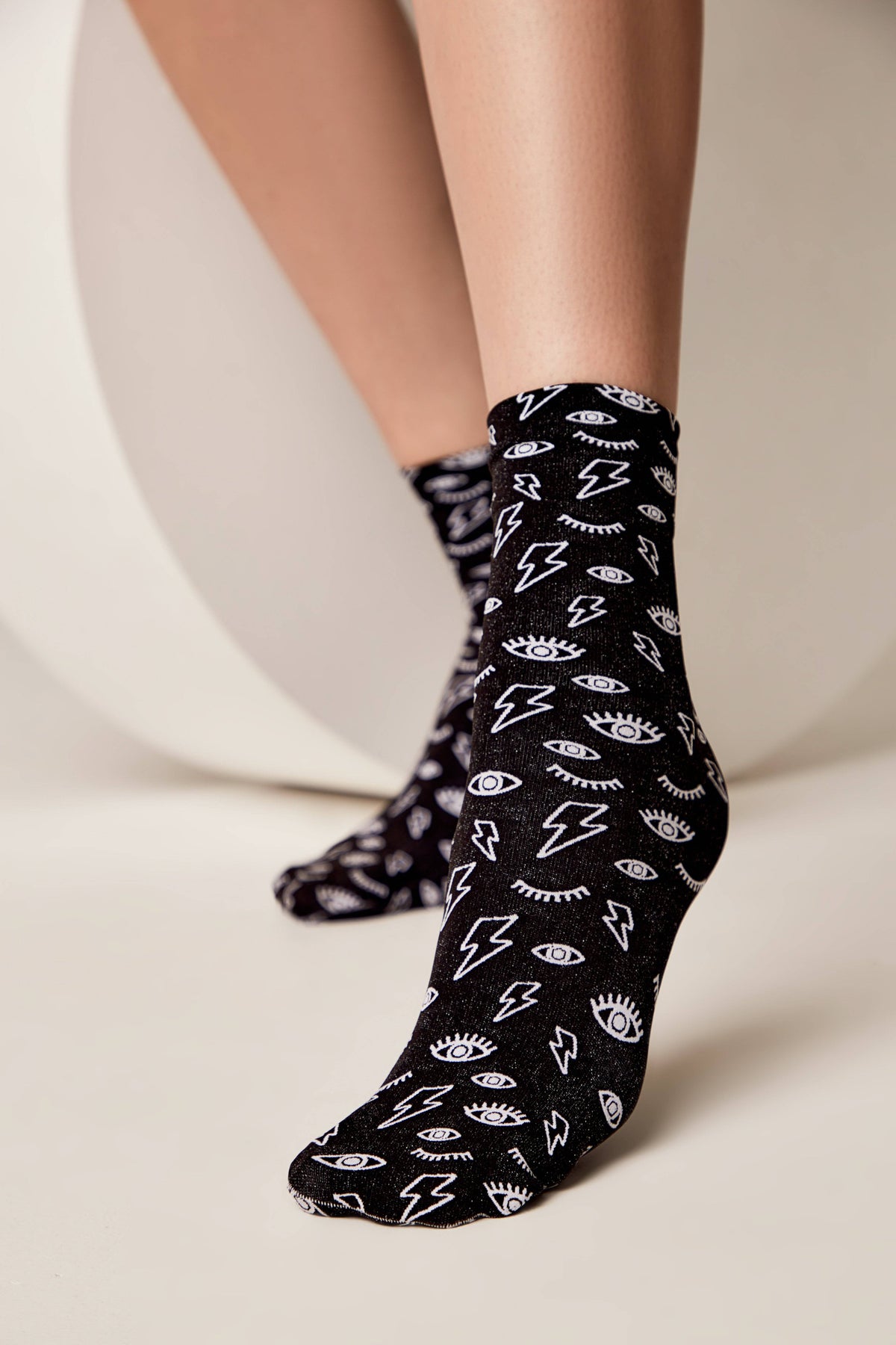 Black crew socks featuring a repeated white pattern of lightning bolts and open eyes worn on a person's feet against a neutral studio background.