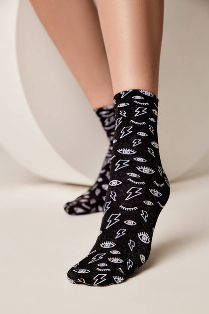 Black crew socks featuring a repeated white pattern of lightning bolts and open eyes worn on a person's feet against a neutral studio background.