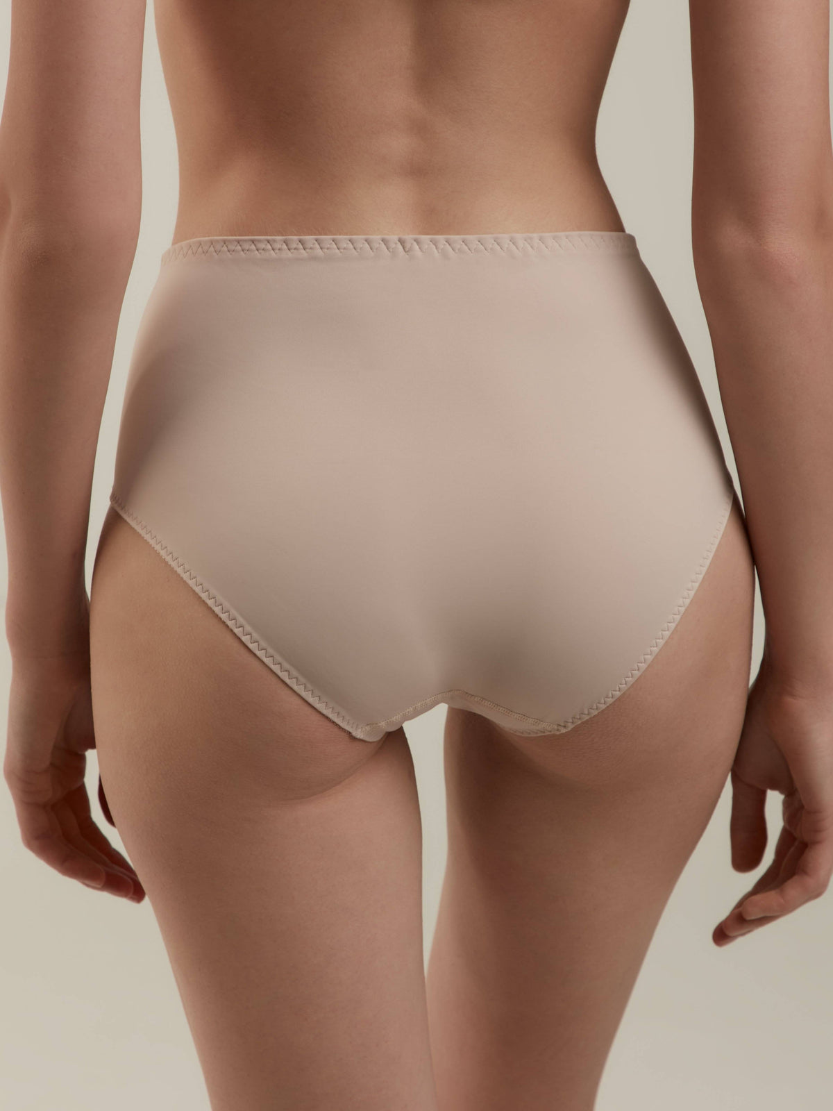 A woman seen from behind wearing beige high-waisted briefs with visible stitching along the waistband and leg openings.