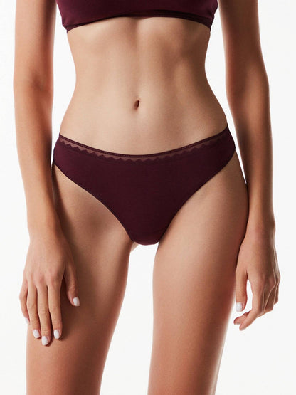 A pair of deep burgundy high-cut bikini-style panties with a delicate geometric patterned trim along the waistband, worn by a person against a plain white background.