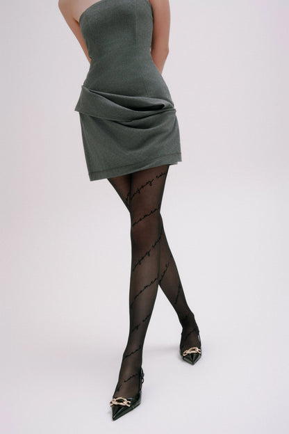 Black sheer tights with repetitive cursive script text detailing, styled with a grey strapless mini dress and black pointed-toe heels with gold-tone hardware.