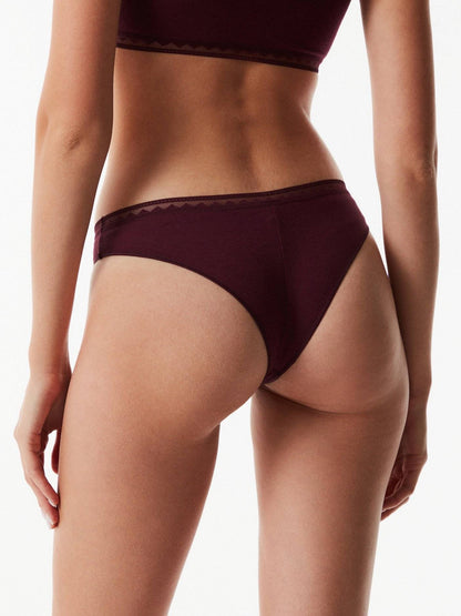 A back view of a person wearing deep burgundy, high-cut bikini-style underwear with a decorative scalloped lace waistband against a plain white background.