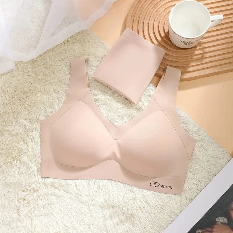 A beige seamless wireless bra is laid flat on a white faux fur rug next to a folded piece of matching fabric, a wooden coaster, and a white mug.