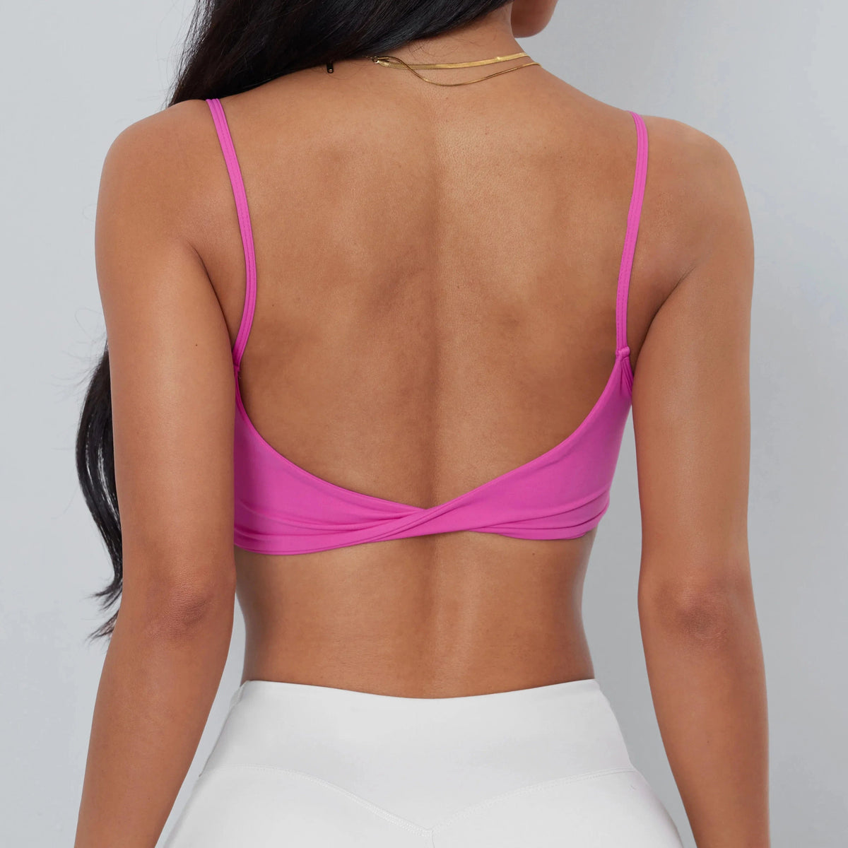 A back view of a person wearing a bright pink cropped camisole with thin straps and a twisted detail at the lower back, paired with white athletic leggings.