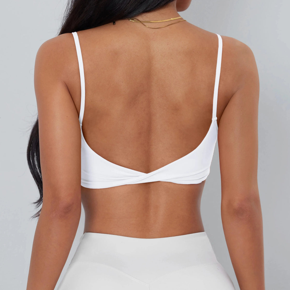 A white sports bra with spaghetti straps and a twisted detail at the center back, worn by a person against a light gray background.