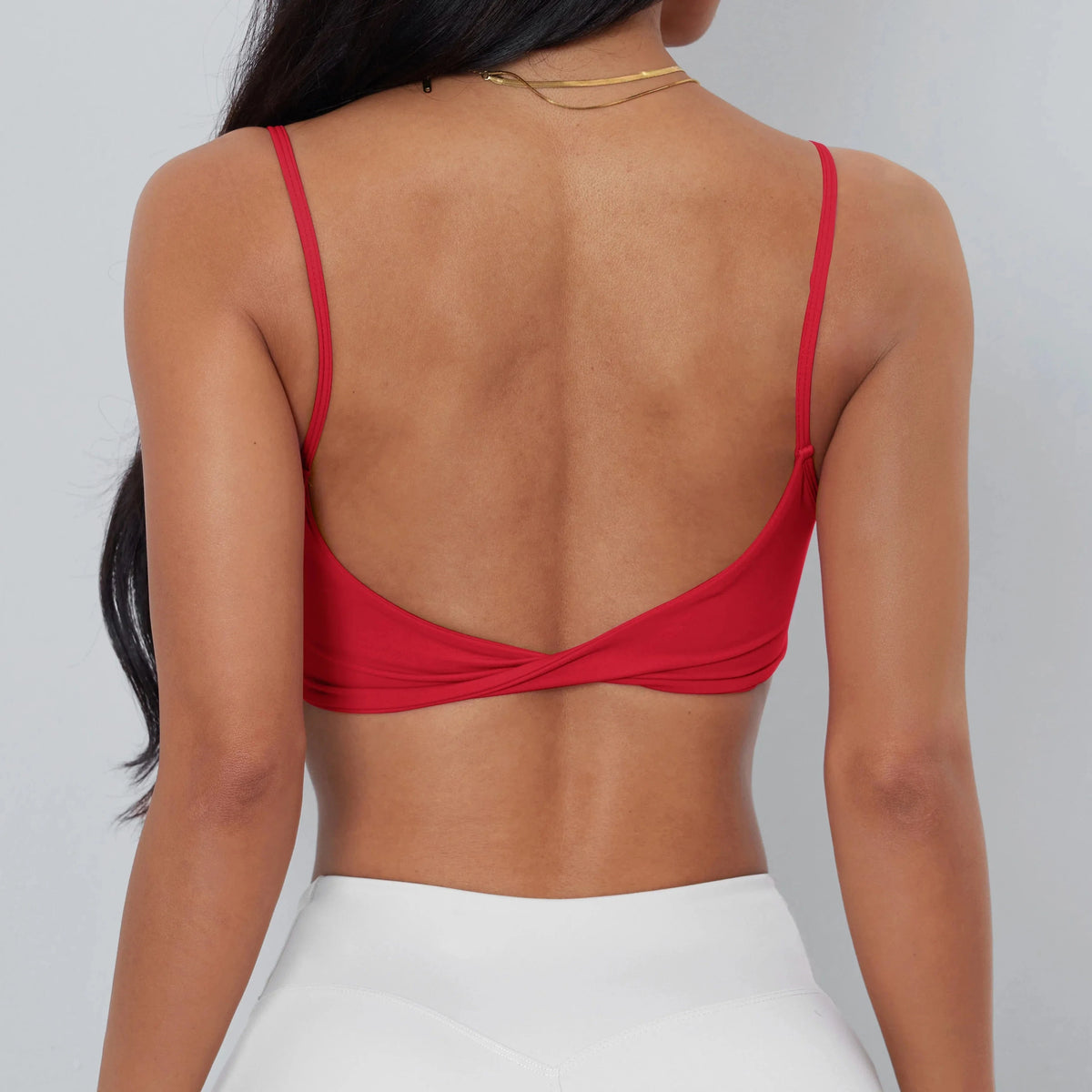 A back view of a person wearing a bright red cropped camisole top featuring thin straps and a twisted hem detail, paired with white athletic leggings.