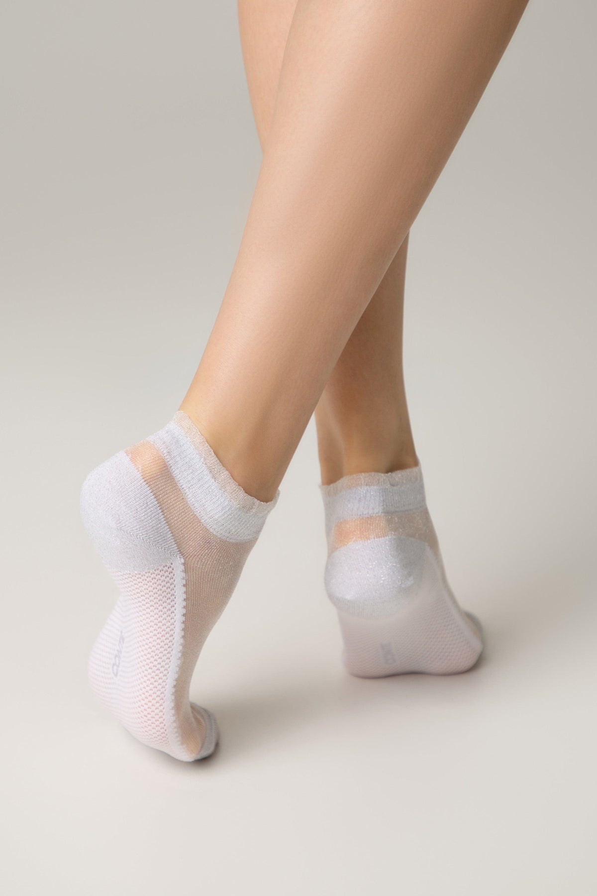 Sheer white low-cut ankle socks with a shimmering finish and reinforced toes and heels worn by a model against a neutral background.