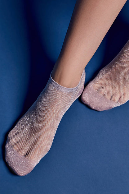 A pair of sheer, shimmering light pink ankle socks with a fine mesh texture worn on a person's feet against a solid blue background.