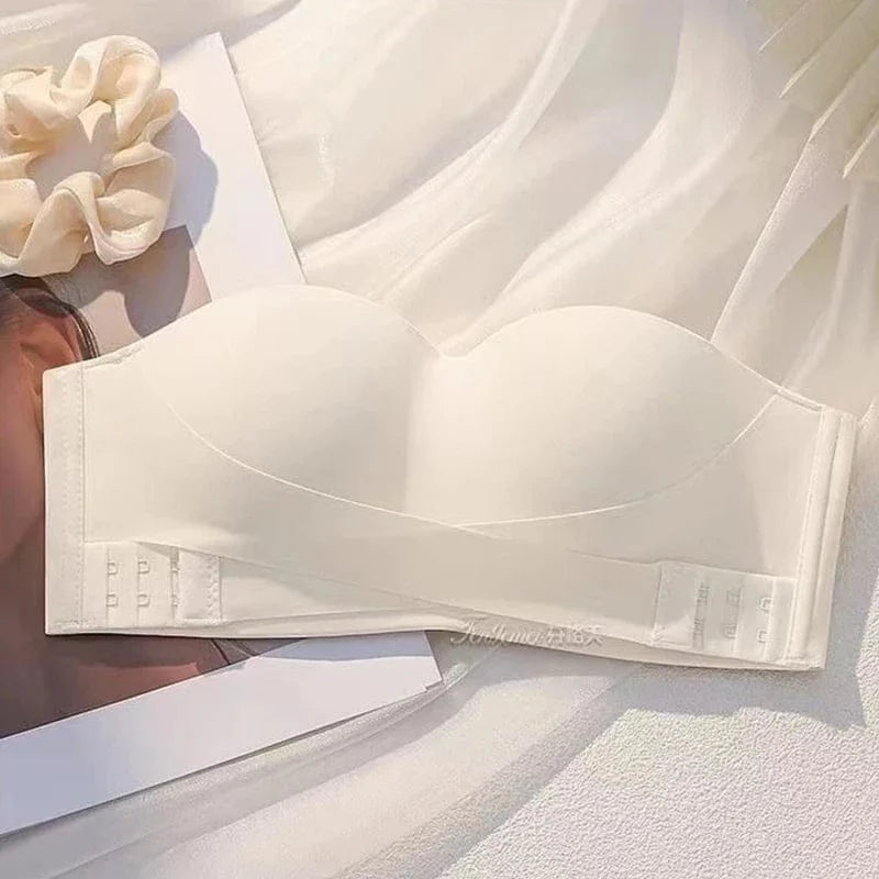 A white strapless bra with a crisscross front design, lying on a white fabric surface next to a cream-colored hair scrunchie.