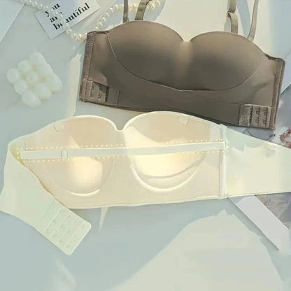 Two strapless bras, one in taupe and one in cream, displayed flat on a white surface alongside pearl strands and decorative square candles.
