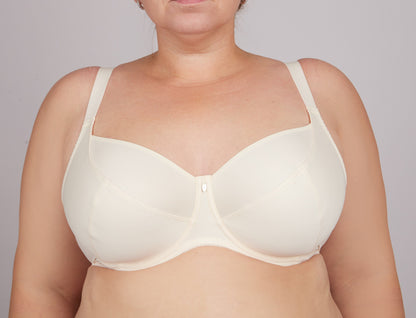 A beige full-coverage underwire bra worn by a person, featuring smooth cups, wide adjustable straps, and a small central charm detail.
