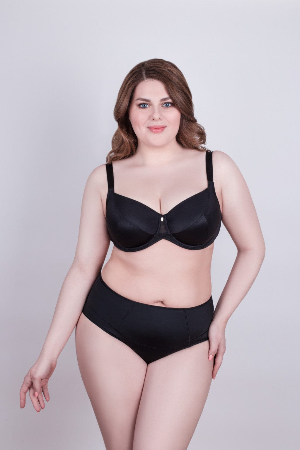 A woman wearing a black matching bra and high-waisted panty set against a plain light gray background.