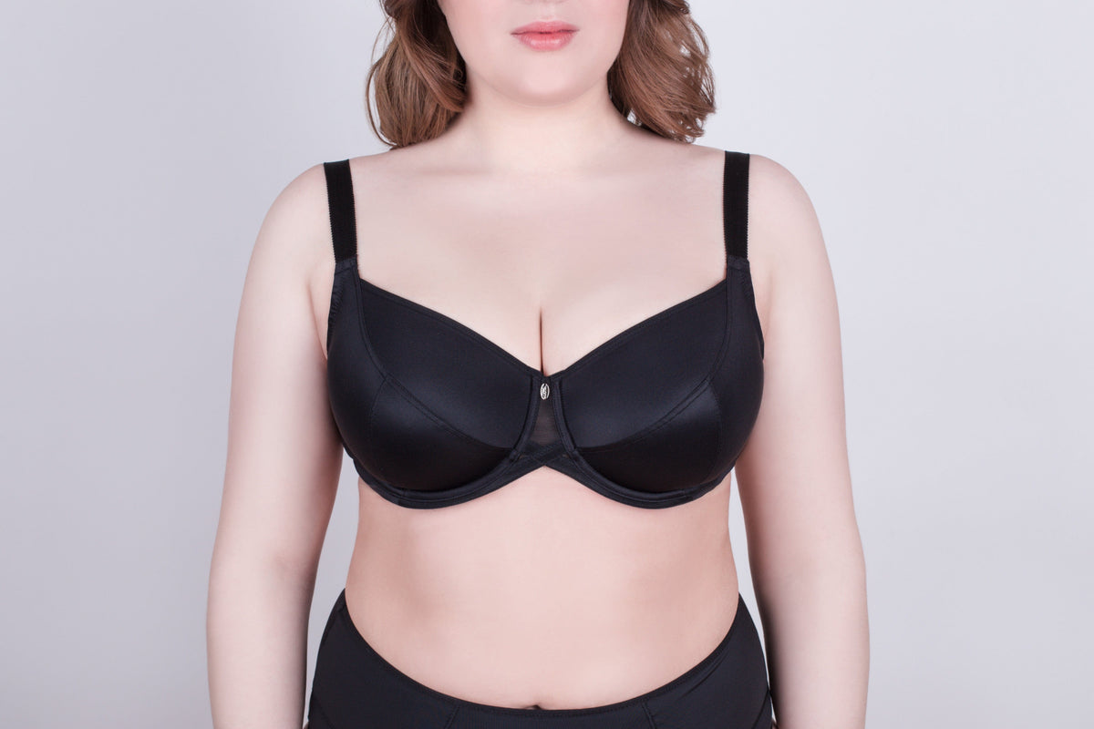 A black balconette bra with wide shoulder straps and a small central charm worn by a person against a plain light gray background.