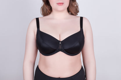 A black balconette bra with wide shoulder straps and a small central charm worn by a person against a plain light gray background.