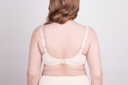 A beige bra shown from the back on a person with fair skin, featuring adjustable shoulder straps and a hook-and-eye closure.