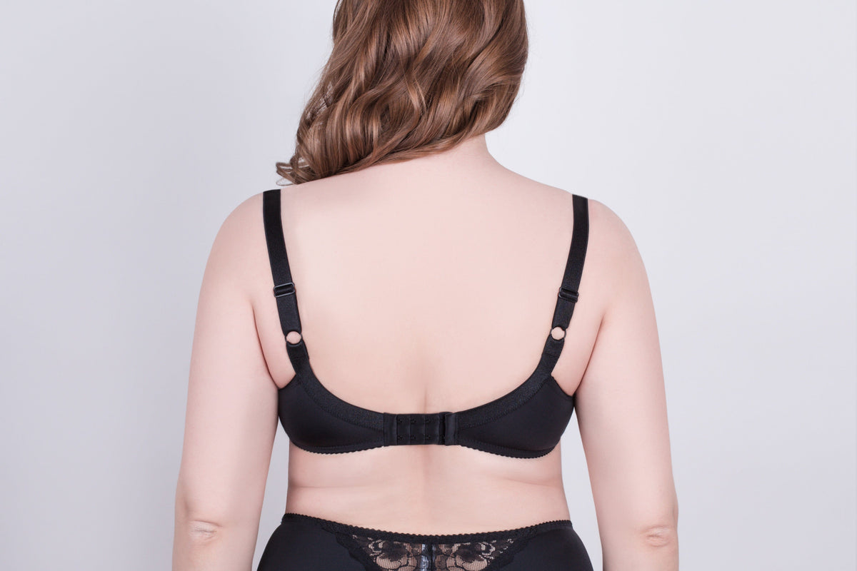 A black bra viewed from the back on a person with long wavy brown hair, featuring adjustable shoulder straps and a hook-and-eye closure.