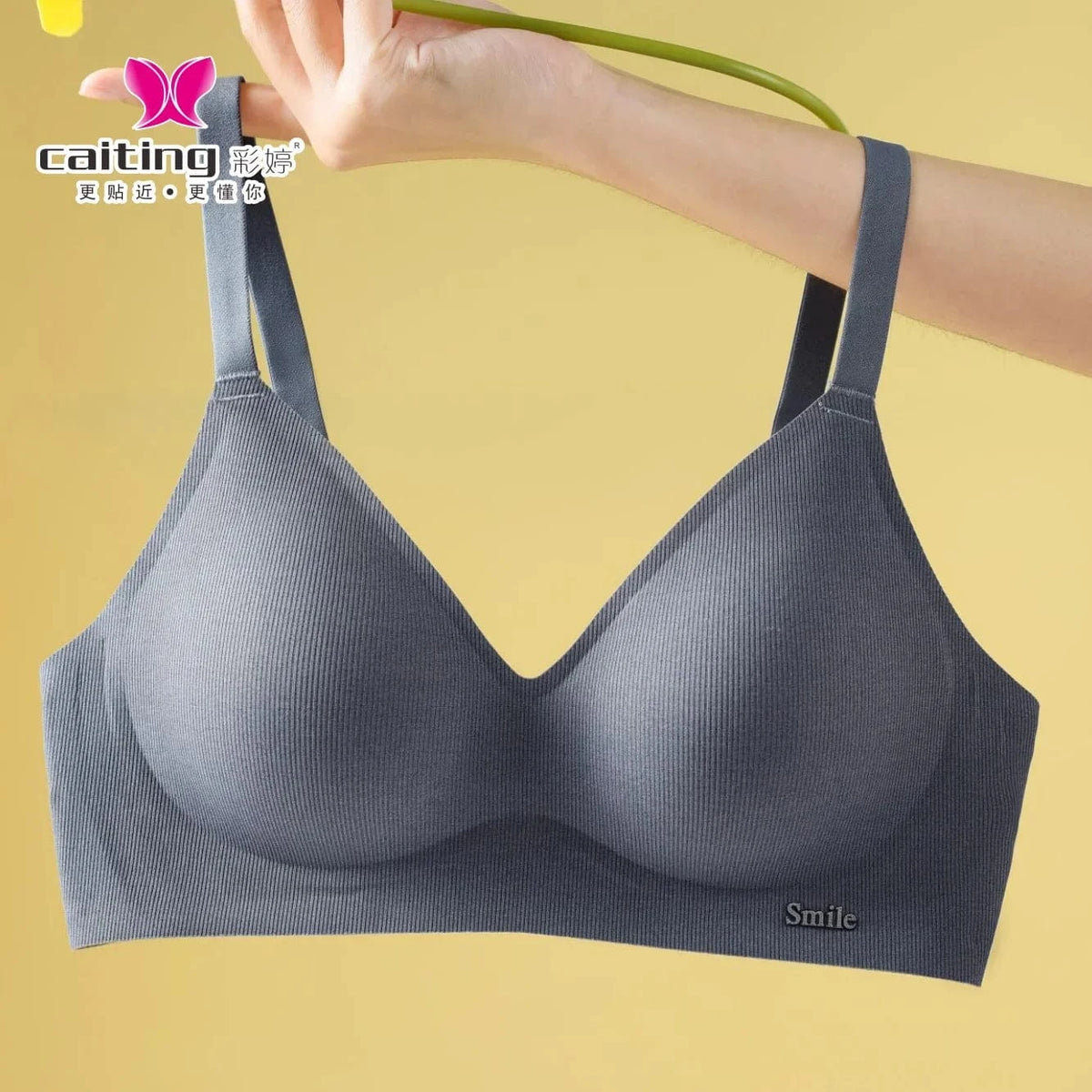 A grey seamless bralette with ribbed texture, wide straps, and a small metal smile accent on the bottom right corner held against a solid yellow background.