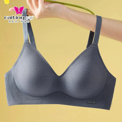 A grey seamless bralette with ribbed texture, wide straps, and a small metal smile accent on the bottom right corner held against a solid yellow background.