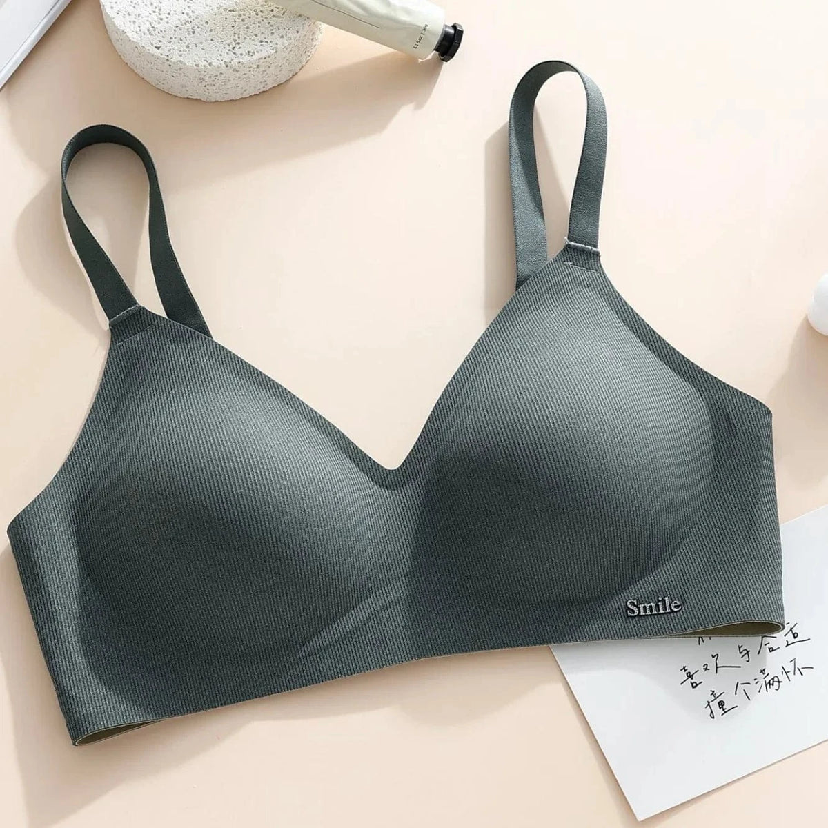 A dark green ribbed bralette with adjustable straps and a small metal "Smile" label on the lower right cup, laid flat on a light beige surface next to a white card and a tube.