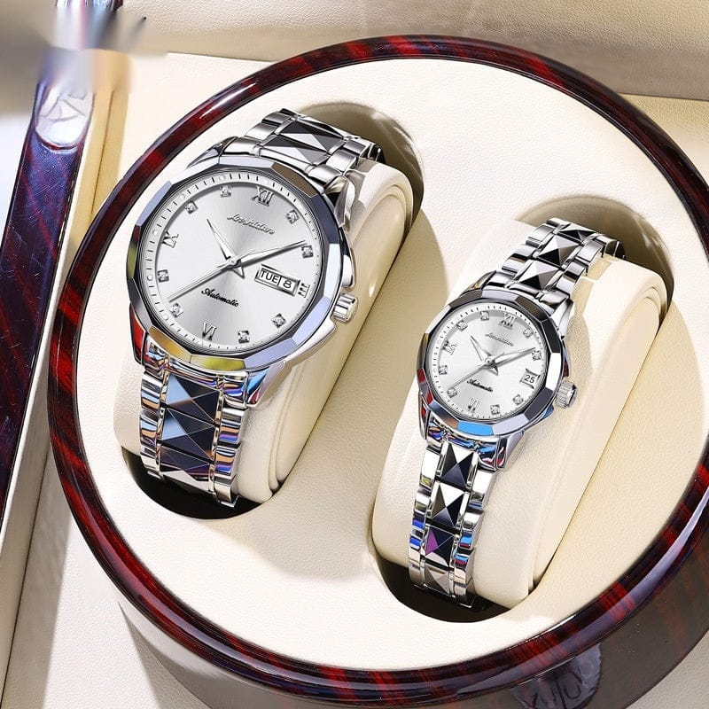 A pair of silver-toned automatic watches with linked geometric bands and white faces featuring crystal hour markers and date displays are presented side by side in a circular wood-grain display box.