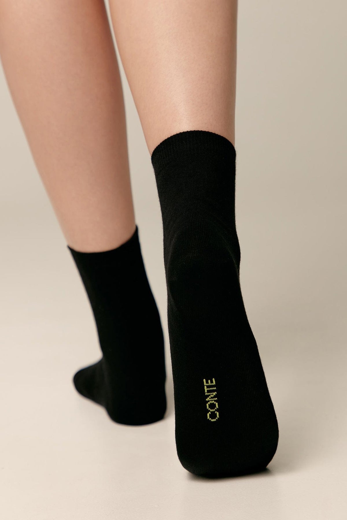 A pair of black ankle-length socks are worn on feet against a neutral background, featuring the word CONTE stitched in light yellow on the bottom of the right sole.
