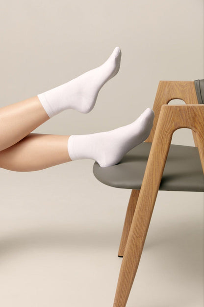 A pair of plain white ankle socks worn on feet resting on the grey seat of a wooden chair.
