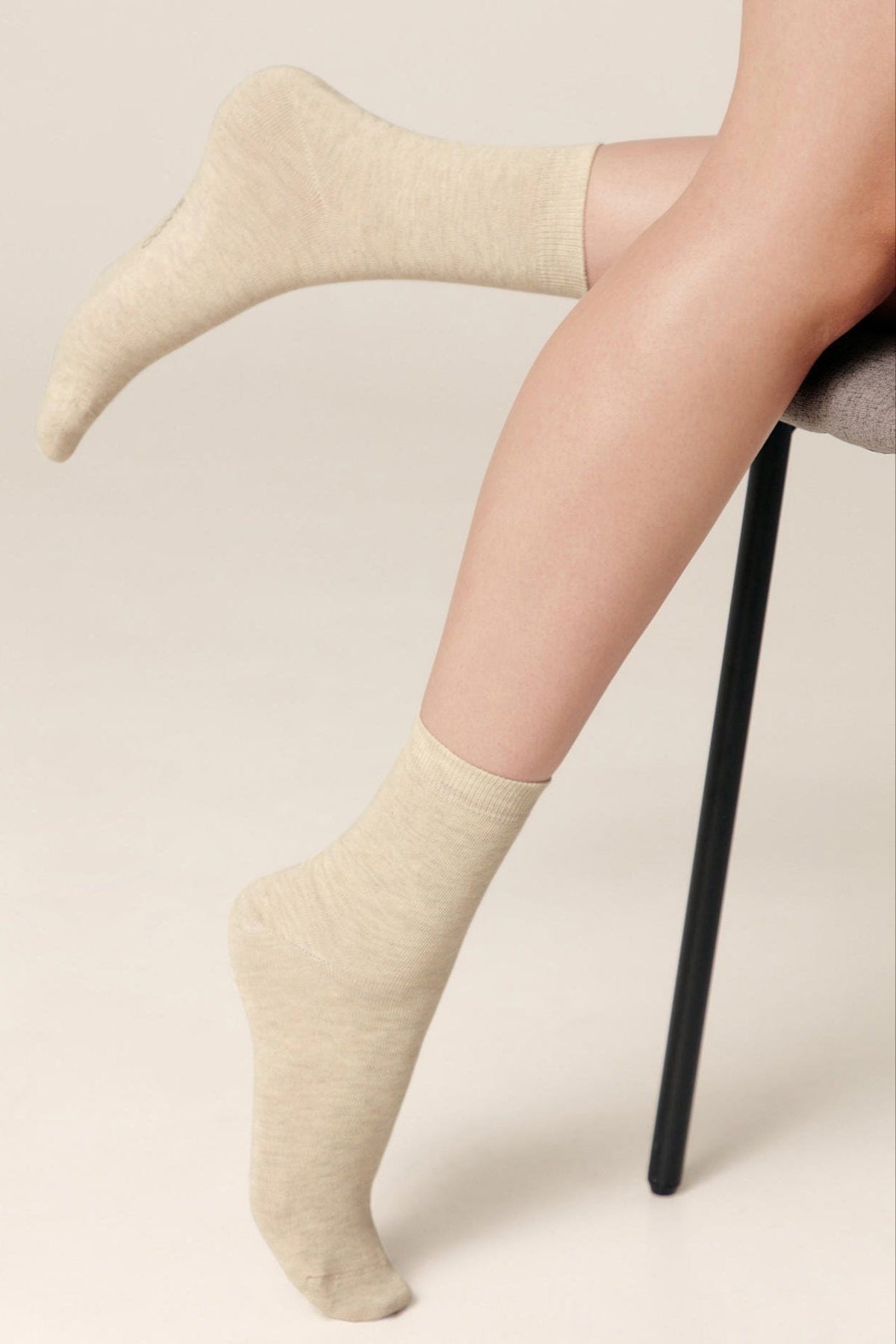 A pair of light beige crew socks worn on a person's feet, showing the ribbed cuff and smooth knit texture while they sit on a grey chair.