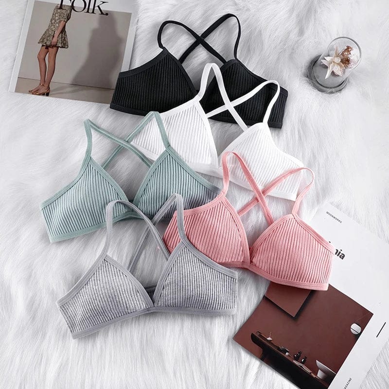 Five ribbed bralettes with crisscross straps are arranged on a white faux fur surface, featuring black, white, light blue, pink, and grey colorways.